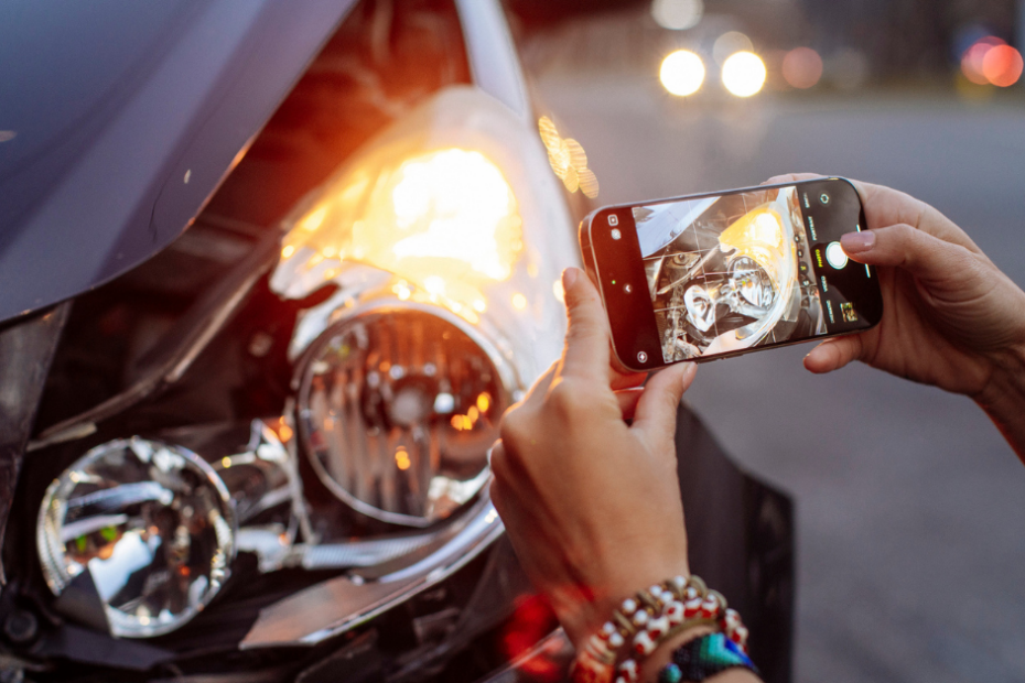 close up image of a person taking a photo of their car's damage after being hit by an uninsured driver. shows camera in hand and broken front headlight
