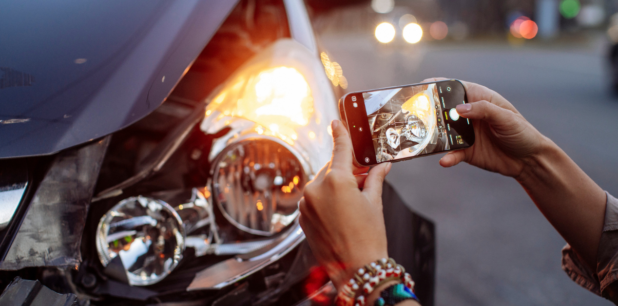 close up image of a person taking a photo of their car's damage after being hit by an uninsured driver. shows camera in hand and broken front headlight